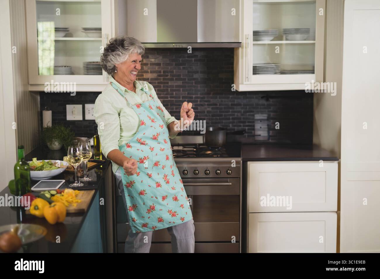 Donna anziana che indossa un grembiule che danzava, tagliava i peperoni sul bancone in una cucina moderna con un'insalata Foto Stock
