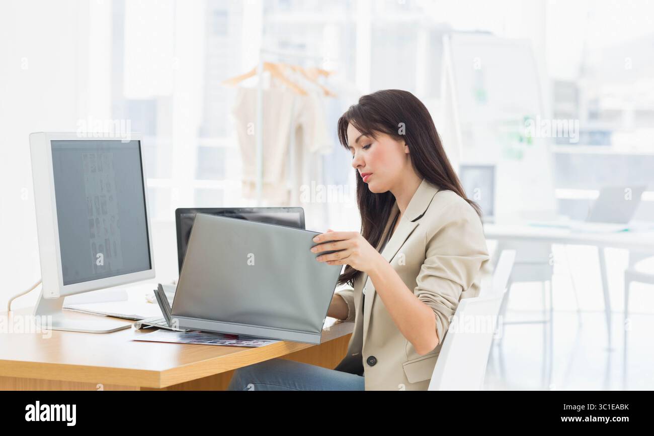 Donna che lavora su un computer portatile con monitor esterno e appendiabiti dietro mentre è seduta alla scrivania dell'ufficio Foto Stock