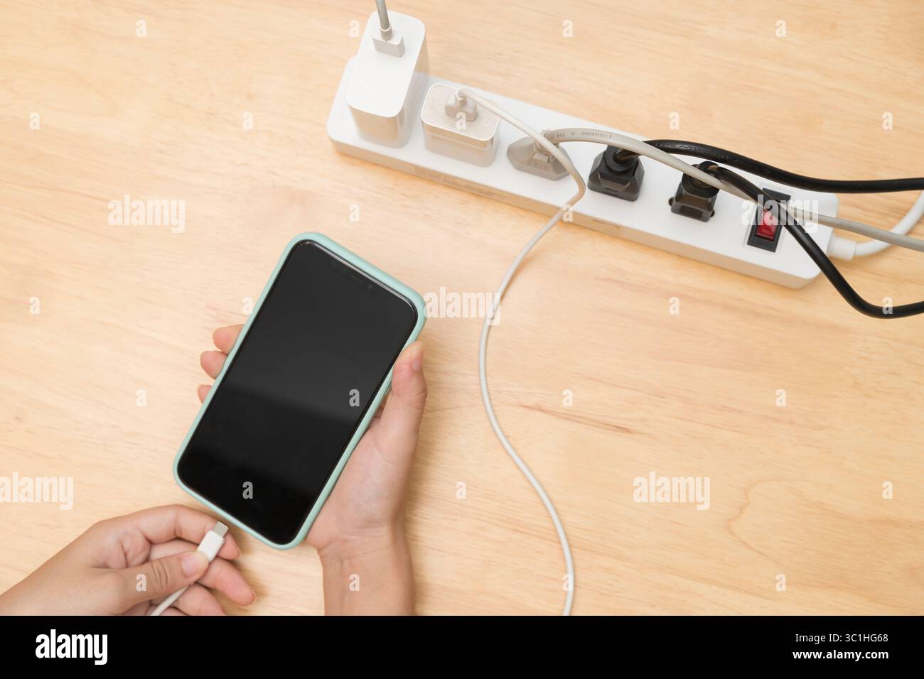 Persona che tiene in mano uno smartphone e inserisce il cavo di ricarica in una presa multipla su una scrivania di legno piena di varie spine e adattatori, come illustrato nella figura Foto Stock