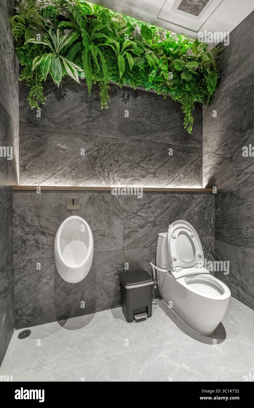 Bagno moderno con WC e orinatoio contro pareti in marmo scuro, con design biofilo con un lussureggiante soffitto di piante artificiali verdi Foto Stock