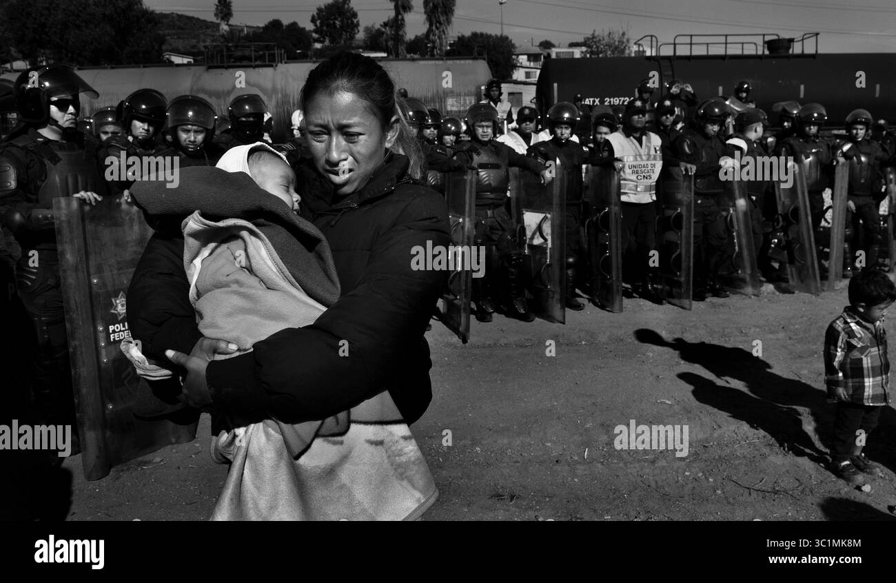 25 novembre 2018 - Tijuana, Messico - ROSA VILLA e il suo bambino ESTEBAN, di 5 mesi, passano davanti alla polizia messicana in attrezzatura antisommossa allontanando i migranti dal confine. Vengono dall'Honduras. La carovana migrante dal campo Centro Deportivo Benito Juarez marcia fino al confine Tijuana-San Diego. Sono stati incontrati da una linea di polizia messicana in attrezzatura antisommossa sul ponte, ma sono riusciti a correre oltre loro, salendo su pareti ripide, attraversando il Rio Tijuana e il terreno difficile, raggiungendo il confine, ma sono stati incontrati a volte da gas lacrimogeni degli agenti statunitensi quando hanno cercato di salire sopra la recinzione e hanno supplicato di essere lasciati entrare. EV Foto Stock