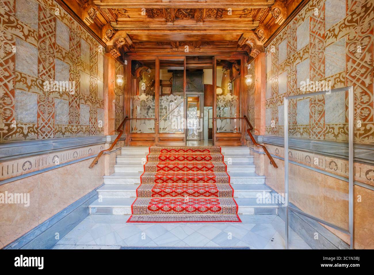 Ingresso al lussuoso edificio di Barcellona con scalinata con moquette rossa. Pareti ornate, lavorazioni in legno dettagliate. Architettura storica spagnola, elegante centro urbano Foto Stock