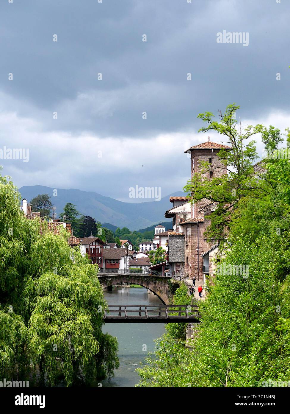 Vista del fiume Nive, del vecchio ponte in pietra e della torre dell'orologio a Saint-Jean-Pied-de-Port, Paesi Baschi francesi, sotto un cielo tempestoso Foto Stock