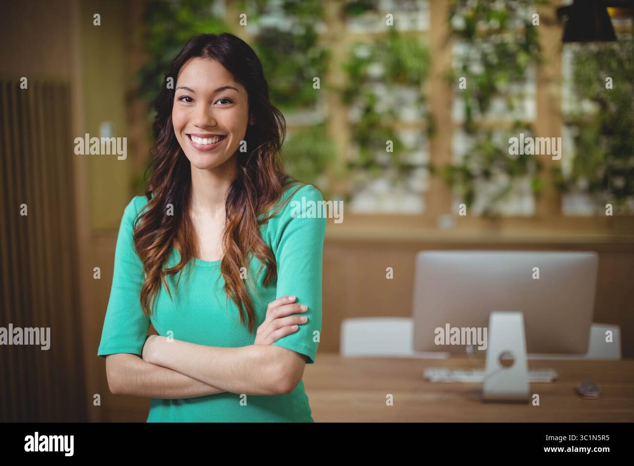 Donna professionista in ufficio sorridente mentre si incrociano le braccia alla scrivania con parete dell'impianto, spazio di copia Foto Stock