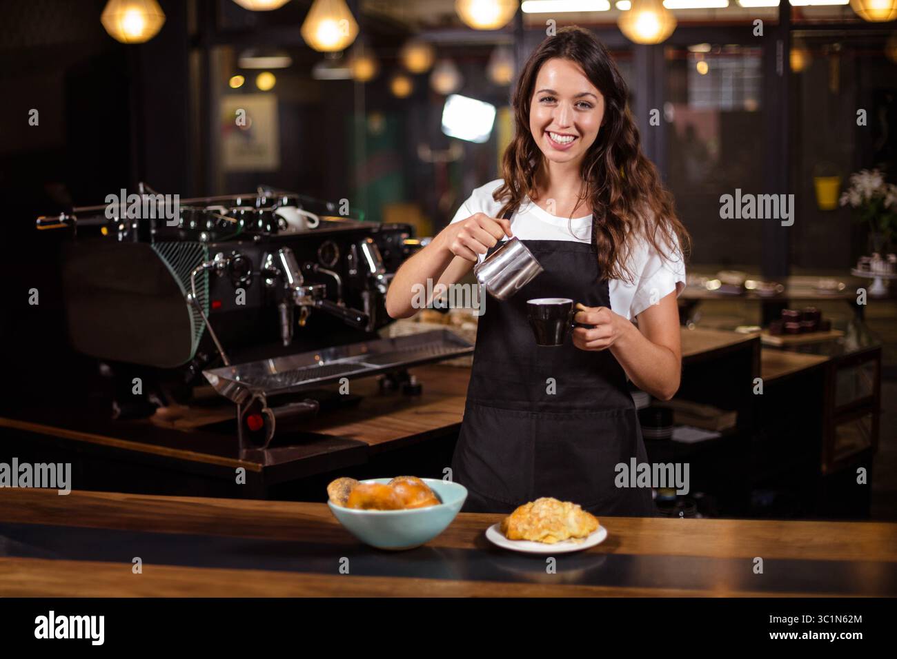 All'interno del caffè, il latte montato che viene versato dalla caraffa metallica nella tazza da caffè su un piatto con croissant Foto Stock