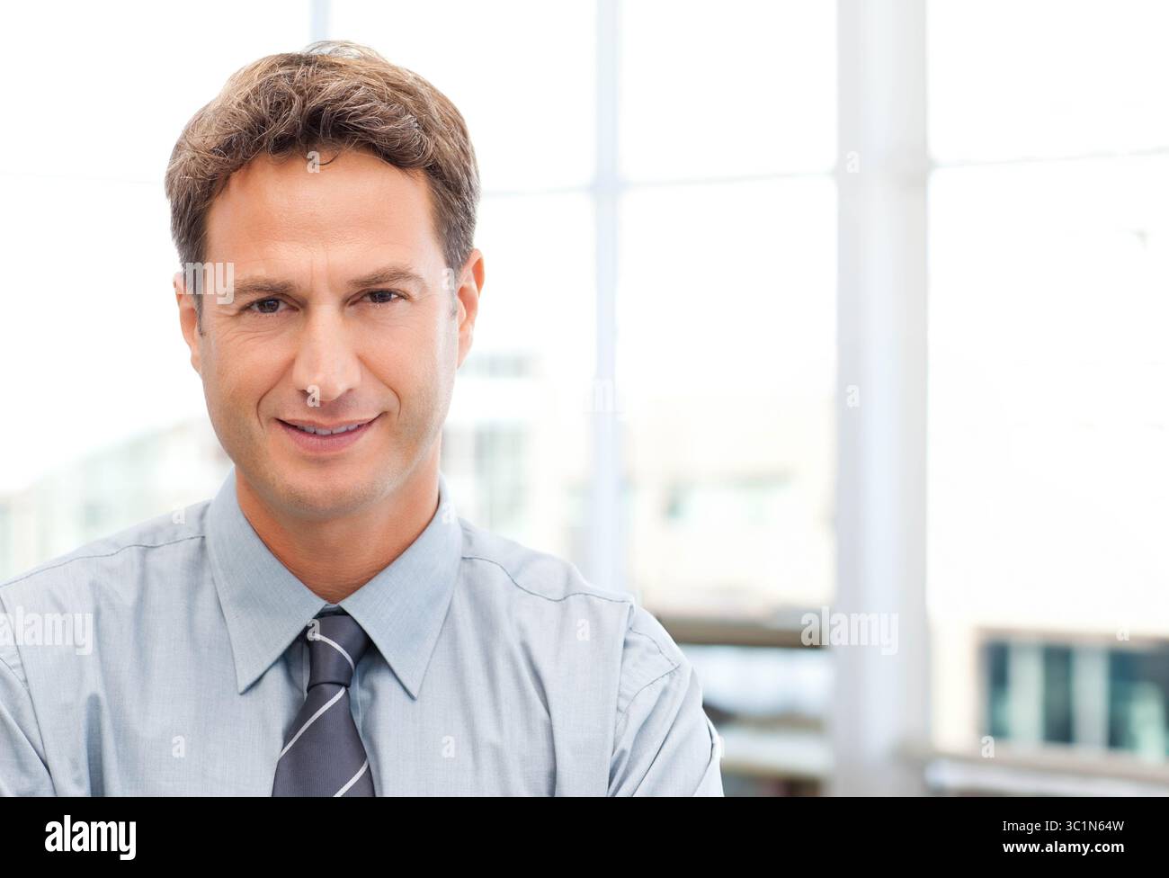 Uomo d'affari che indossa una camicia azzurra con cravatta a righe seduto in ufficio vicino a grandi finestre in vetro Foto Stock