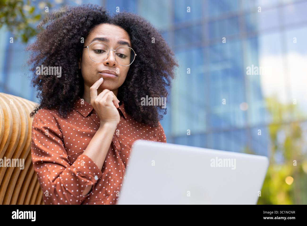Una donna premurosa che indossa occhiali e guarda un notebook all'aperto, riflettendo su una decisione. Foto Stock