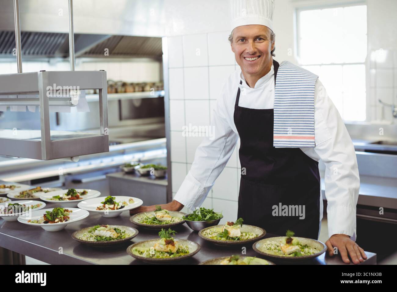 Chef maschile che indossa l'uniforme per preparare i piatti al tavolo sotto le lampade termiche in una cucina affollata, copia spazio Foto Stock