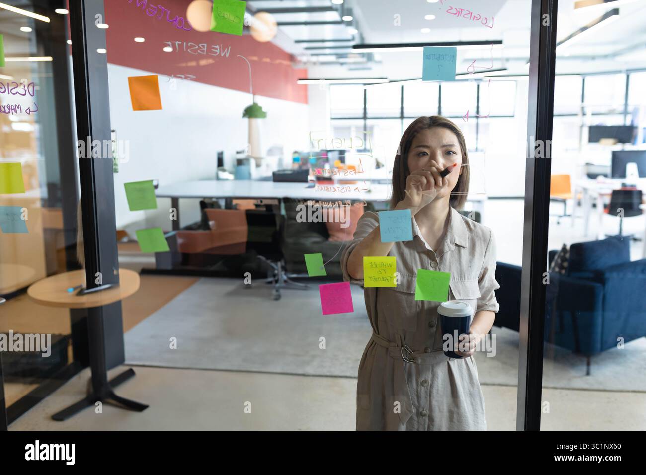 Donna asiatica che scrive su una parete di vetro con note appiccicose mentre tiene una tazza di caffè in un ufficio moderno Foto Stock