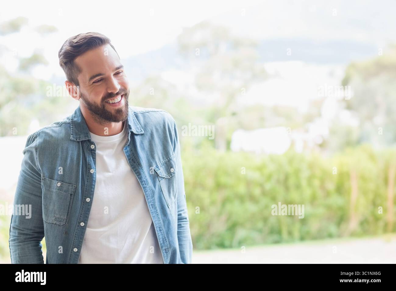 Uomo medio adulto in piedi in giardino con camicia in denim e t-shirt bianca sotto la luce del sole, spazio per copiare Foto Stock