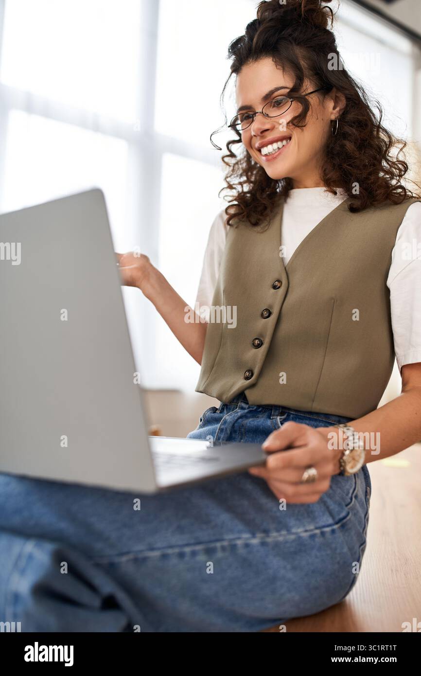 Una giovane donna con i capelli ricci si impegna a lavorare in remoto dal suo moderno appartamento, utilizzando il suo laptop. Foto Stock