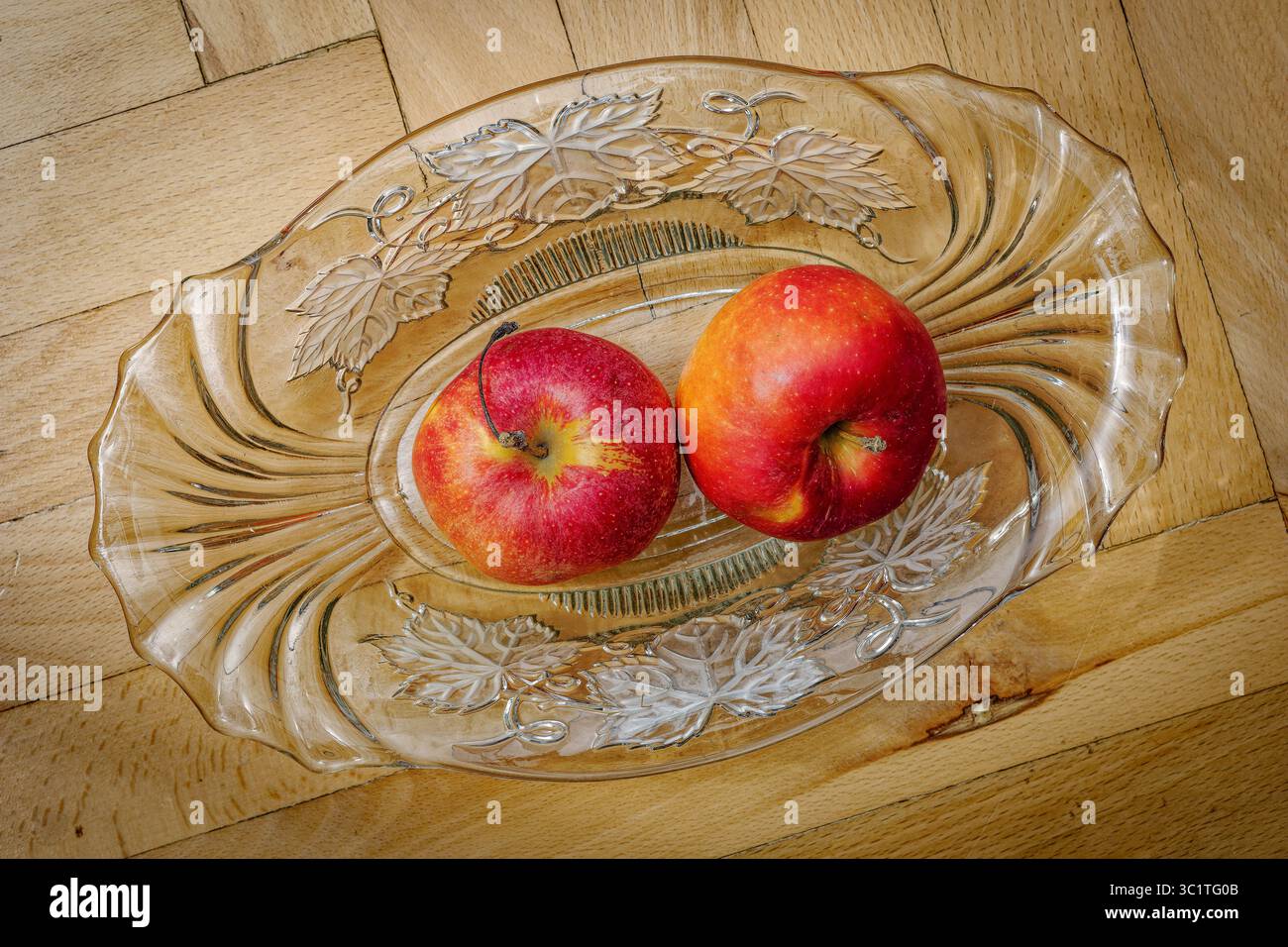 Due mele rosse identiche poggiano su un piatto di vetro trasparente di forma ovale. Il piatto presenta un motivo decorativo di foglie stilizzate lungo i bordi. L'appl Foto Stock