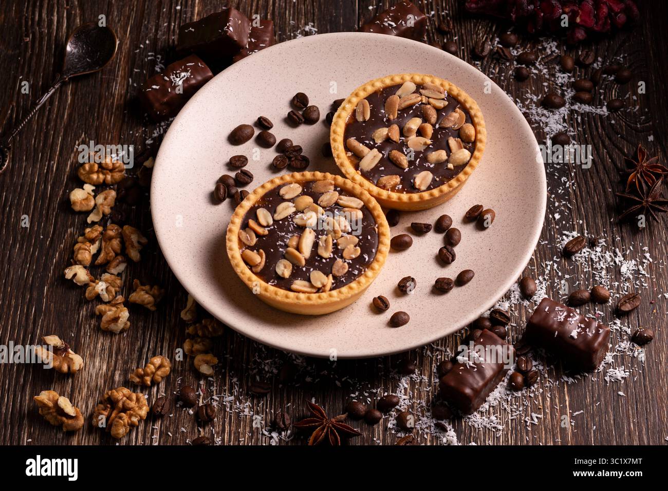 Due crostate di caffè al cioccolato si trovano su un piatto beige, circondate da chicchi di caffè, noci e pezzi di cioccolato. Le crostate sono ricoperte di cioccolato fondente Foto Stock