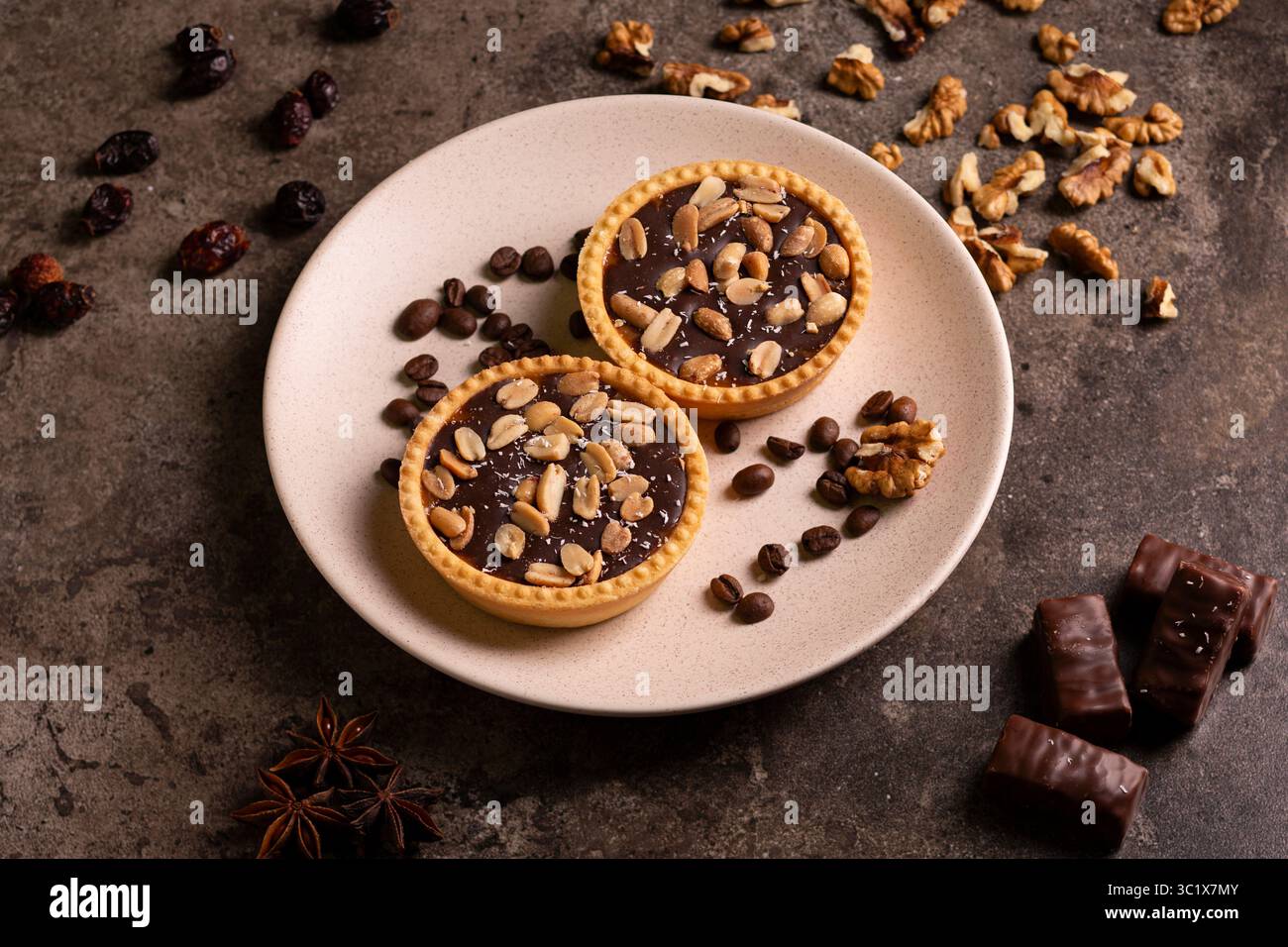 Due crostate di cioccolato fondente si trovano su un piatto beige, guarnite con noci, chicchi di caffè e ciliegie secche. Accanto al PL sono collocate alcune barrette di cioccolato Foto Stock