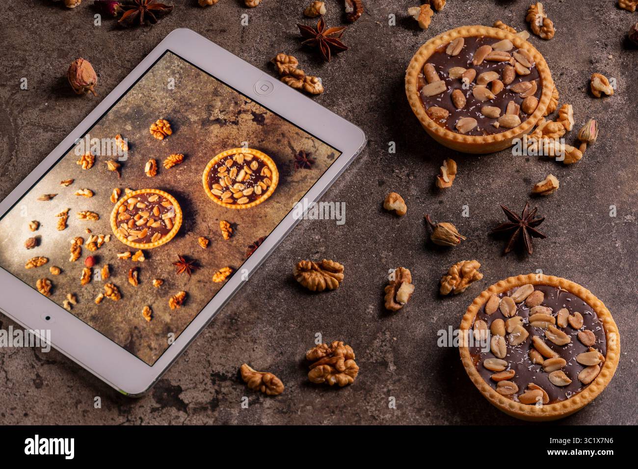 Una tavoletta mostra una fotografia ravvicinata di tre crostate di cioccolato pecan disposte su una superficie di pietra grigio scuro. Sono aggiunte sparse intorno al tablet Foto Stock