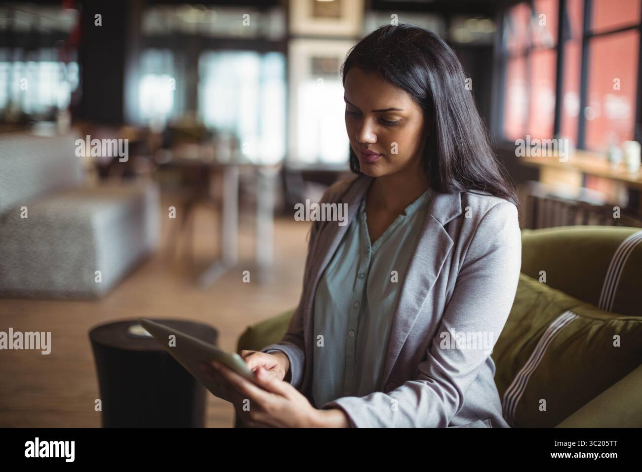 Donna asiatica che indossa abiti da lavoro seduti su una sedia verde oliva tenendo un tablet nella caffetteria lounge Foto Stock