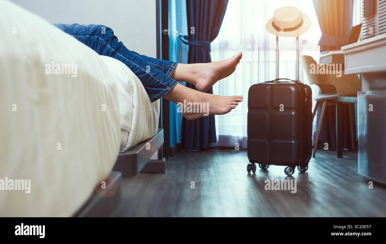 una donna felice e felice viaggiatrice solitaria che si toglie le scarpe, sdraiata su un letto nella camera da letto dell'hotel dopo il check-in con valigia in camera, riposandosi Foto Stock