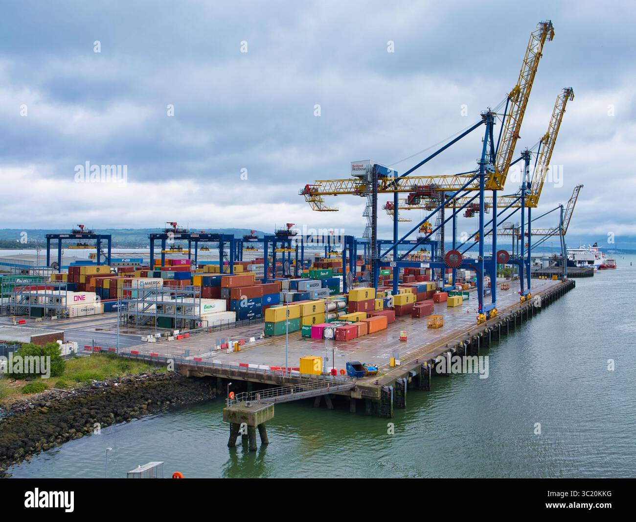 Belfast, Regno Unito - 15 giugno 2025: Grandi gru e container impilati costeggiano i vivaci moli del porto di Belfast sotto un cielo nuvoloso. Foto Stock