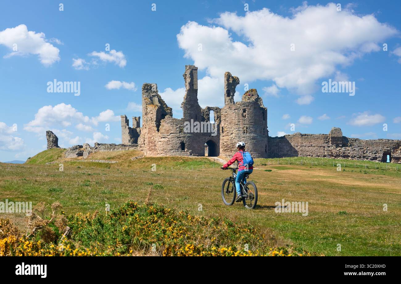 Bella donna anziana che pedala con la sua mountain bike elettrica al castello di Dunstanburgh nel nord-est dell'Inghilterra, Regno Unito Foto Stock