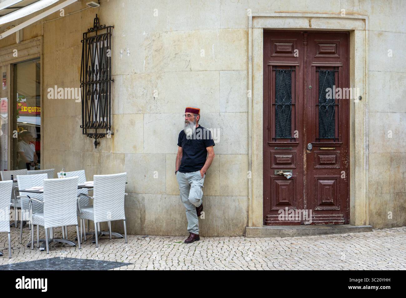 Uomo in piedi contro un muro di pietra vicino a una porta di legno e a un'area salotto all'aperto in una strada pittoresca. Foto Stock