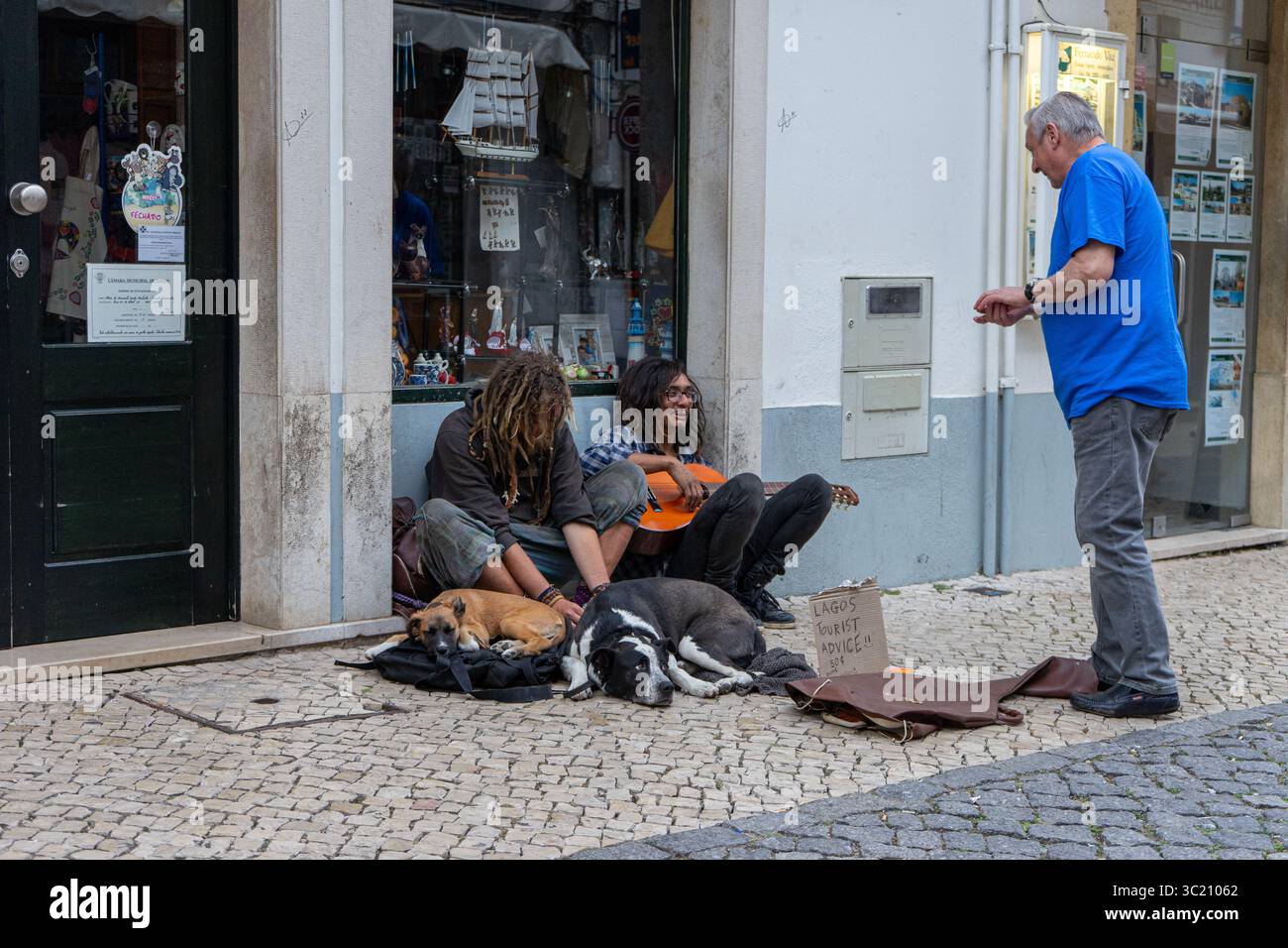 Scena di strada con un uomo in piedi e due persone sedute con un cane su un marciapiede acciottolato di fronte a un negozio. Foto Stock