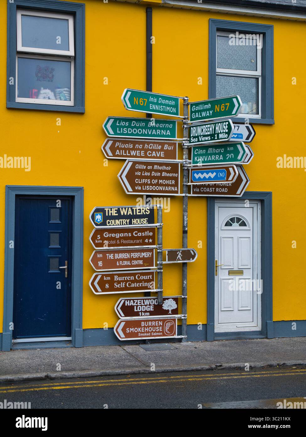 Ballyvaughan, Irlanda - 12 giugno 2025: Una vivace segnaletica piena di direzioni diverse si erge contro un muro giallo brillante a Ballyvaughan, Irlanda. Foto Stock