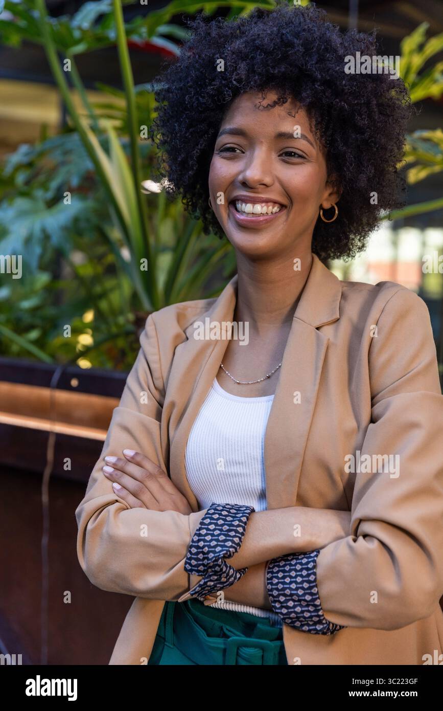 Donna afroamericana che indossa un blazer beige appoggiata su una sporgenza di legno accanto a piante in vaso nel patio del caffè Foto Stock