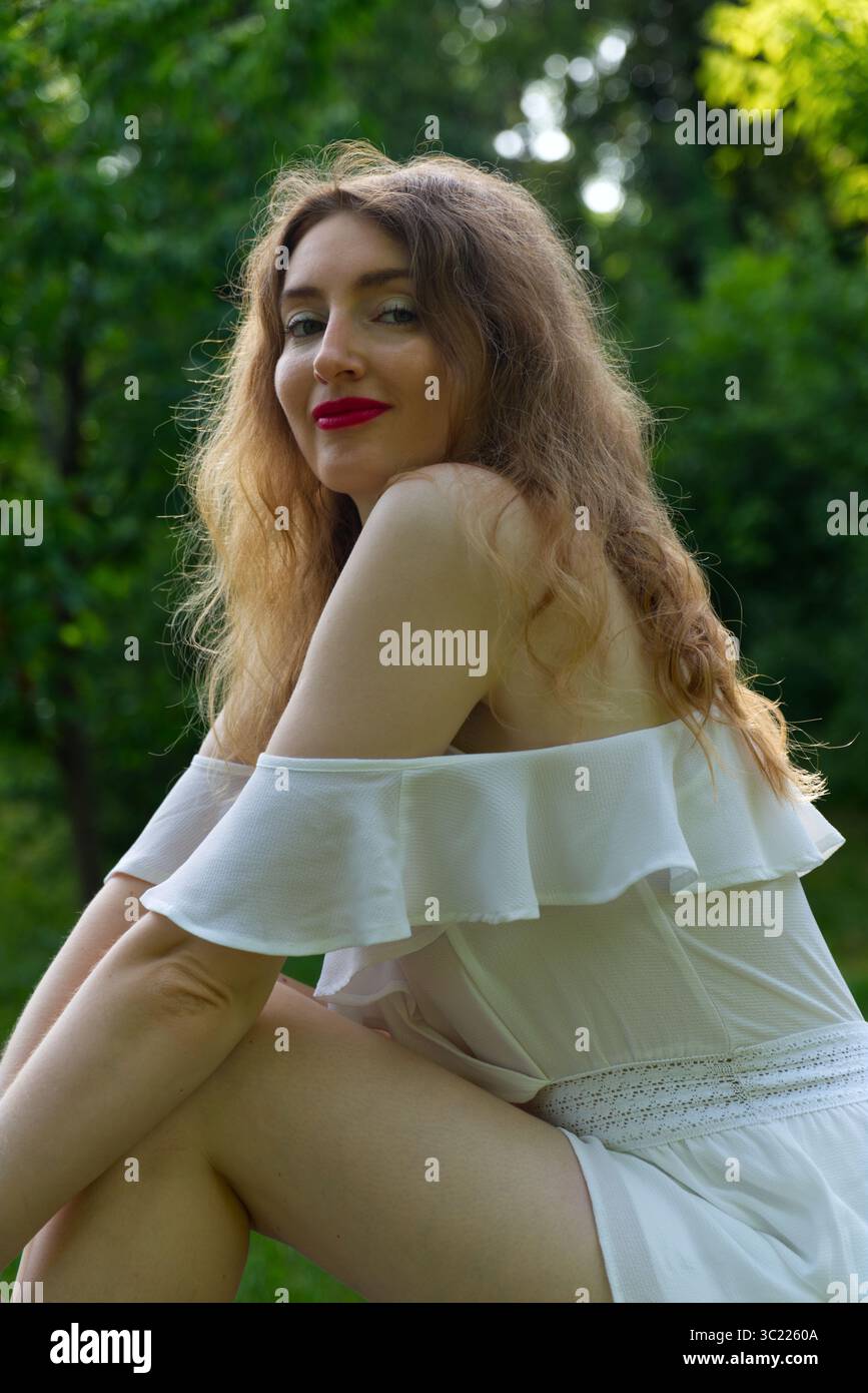 Una donna con i capelli lunghi e ondulati e il rossetto rosso sorride mentre indossa un abito bianco a spalla in un ambiente naturale e verde all'aperto. Foto Stock