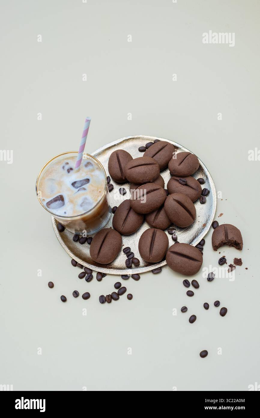 Bevanda a base di caffè ghiacciato con cannuccia colorata accanto a un piatto d'argento di biscotti al cioccolato a forma di chicchi di caffè, creando una scena deliziosa e invitante Foto Stock