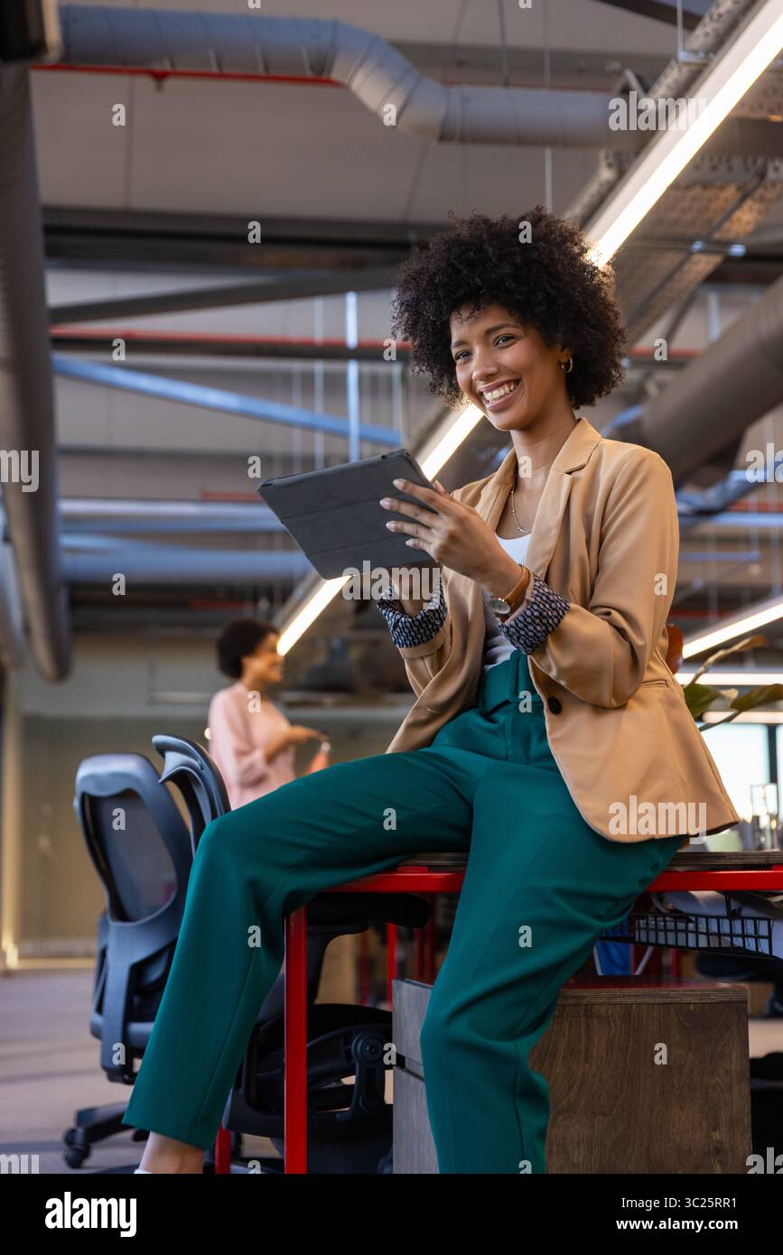 Colleghi di sesso femminile che collaborano su un tablet mentre si siedono su un tavolo rosso alto in un ufficio aperto Foto Stock