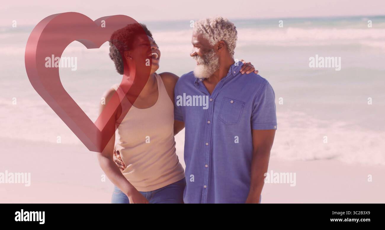 Donna che cammina e uomo maturo con braccio in braccio sorridente lungo la costa sabbiosa, con rivestimento rosso del cuore Foto Stock