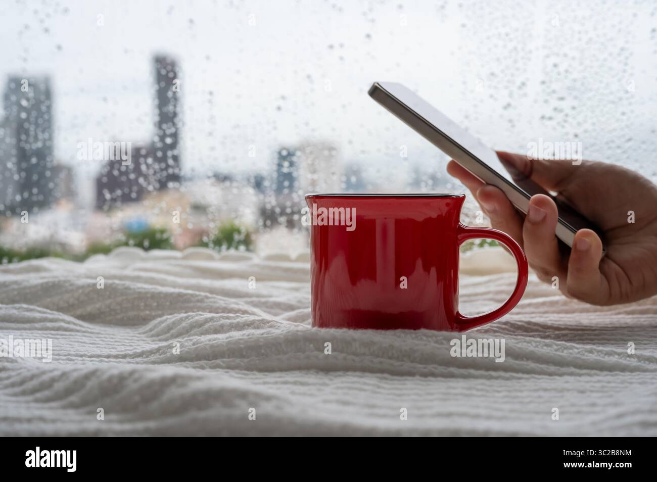 Una tazza di caffè rossa si mette in coperta quando la pioggia cade sul finestrino con lo smartphone che tiene in mano. Soggiorno a casa e relax. Foto Stock