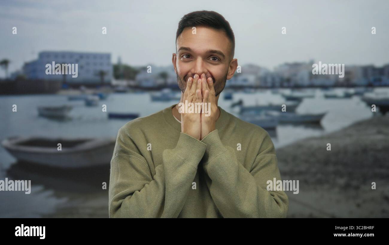 Giovane ispanico che copre la bocca a sorpresa contro il lungomare con barche sullo sfondo, mostrando una candida reazione all'aperto. Foto Stock