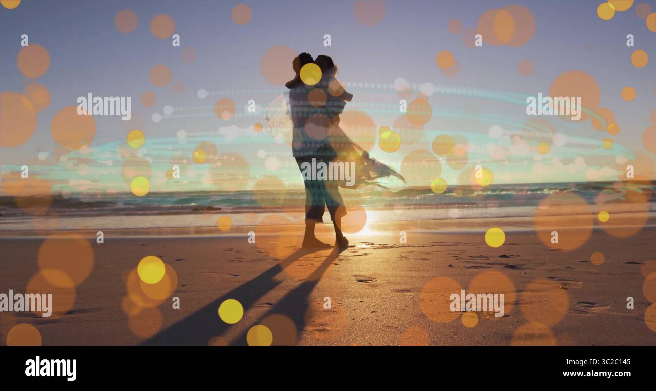 Abbracciare la coppia sulla spiaggia di sabbia sul bordo dell'acqua durante il tramonto, indossando un abito che scorre e una camicia casual Foto Stock