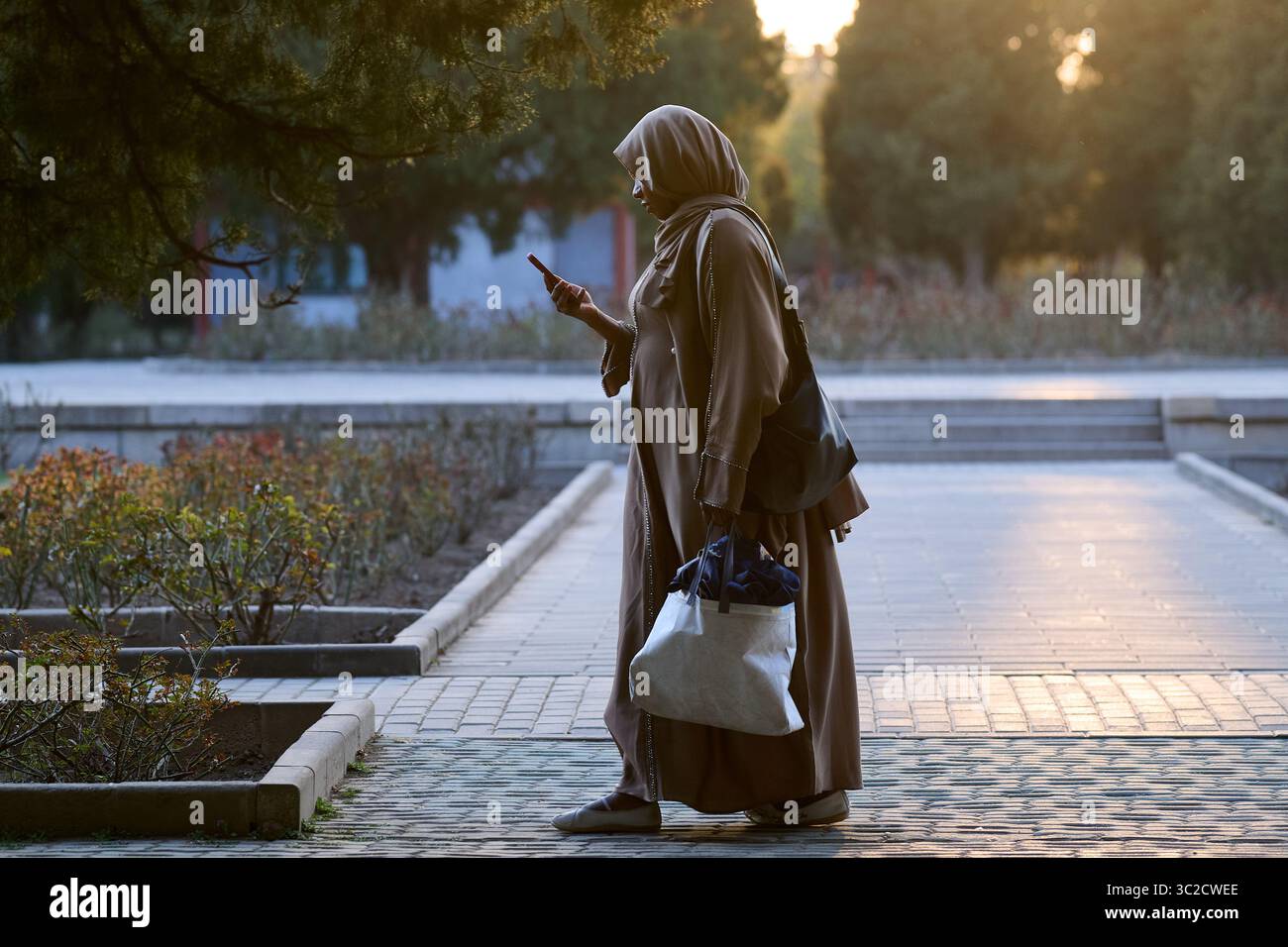 Una donna in abaya marrone cammina accanto a una fontana al tramonto nel parco Foto Stock