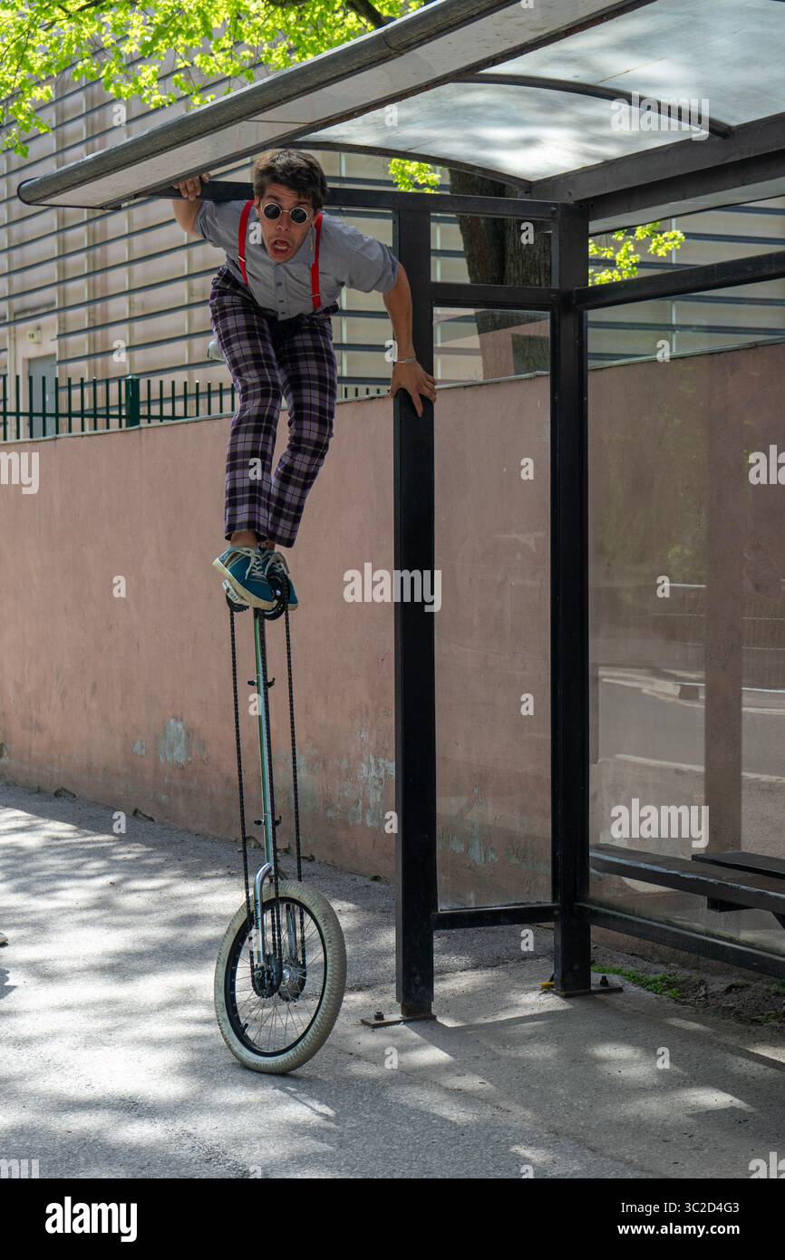 Una persona che guida abilmente un'alta bicicletta in una fermata dell'autobus al riparo in un ambiente urbano, indossando un abbigliamento casual con bretelle e un cappello. Foto Stock