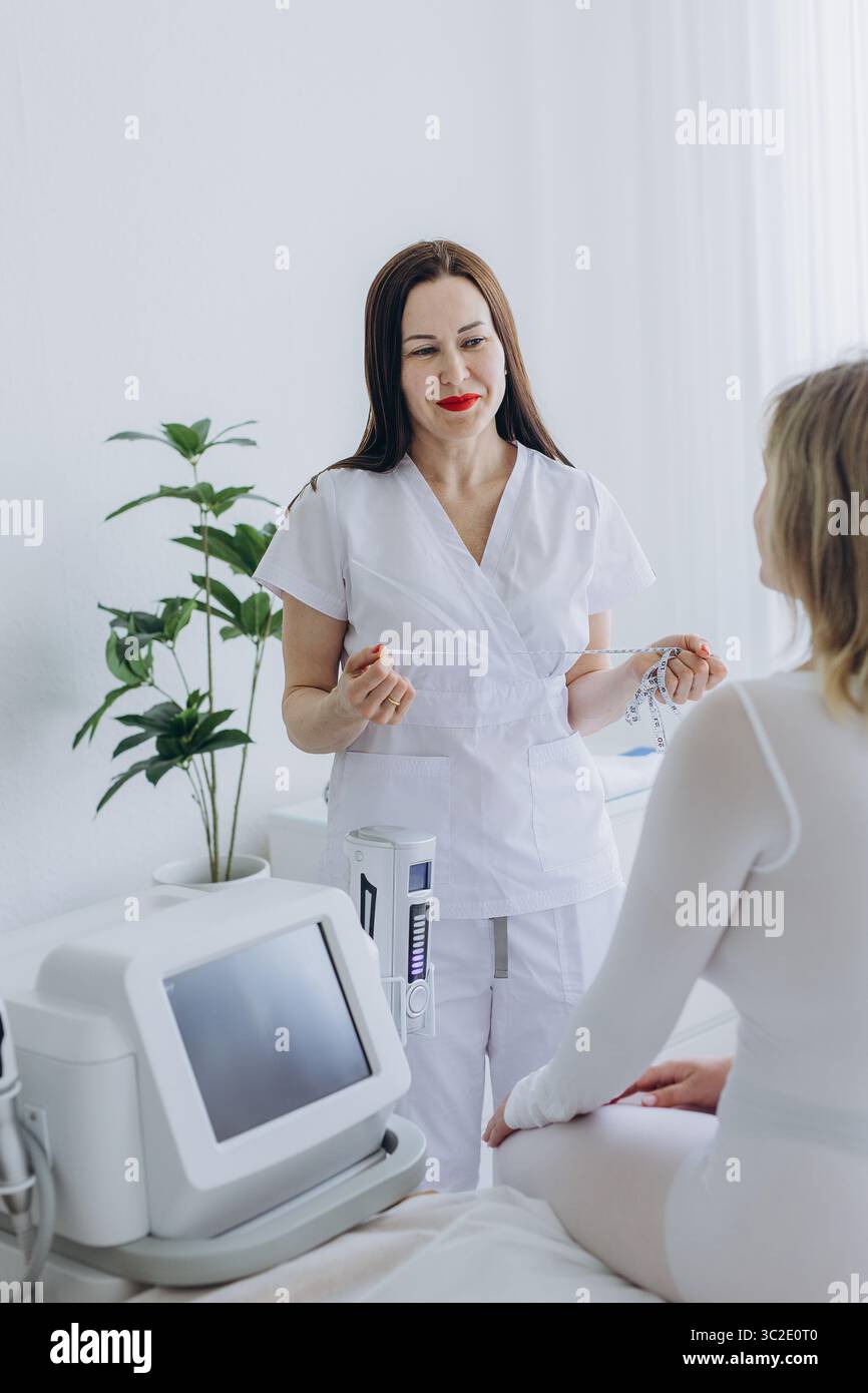 Cosmetologa sorridente con metro a nastro consultando cliente donna prima di contornare il corpo o trattamento anti-cellulite in un moderno centro di bellezza. Alta qu Foto Stock