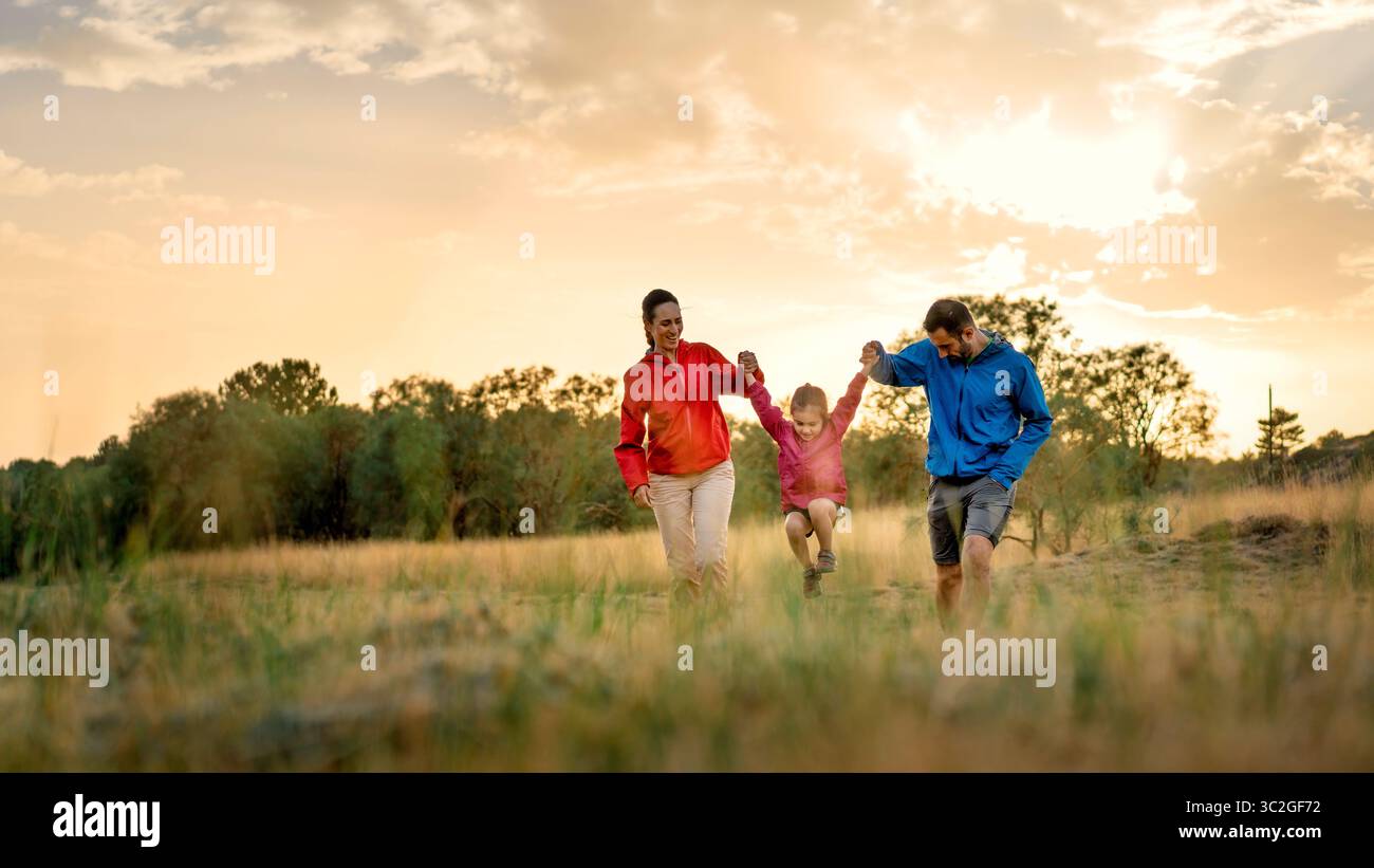 Una foto panoramica cinematografica in 16:9 di una famiglia felice che fa oscillare la figlia in un campo erboso al tramonto, perfetta per un banner o un'intestazione di un sito Web. Foto Stock