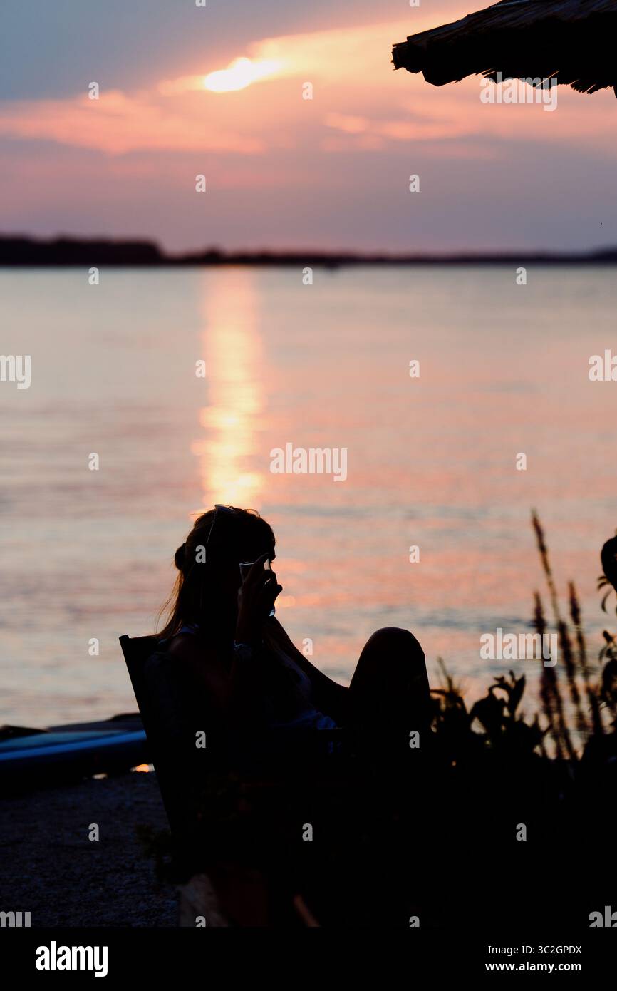 Silhouette di donna irriconoscibile seduta da sola a un tavolo in riva al mare. Bicchiere di birra sul tavolo. Sera d'estate. Fazana, Croazia - circa 2025 Foto Stock