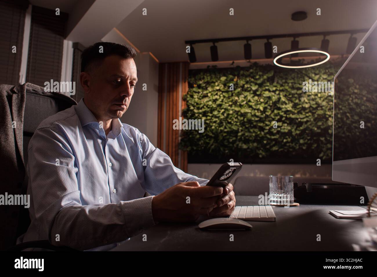Uomo d'affari con una camicia leggera, tastiera e telefono in un ufficio elegante e buio. Luce da sera, spazio di lavoro elegante, concentrazione sulle attività professionali. Foto Stock