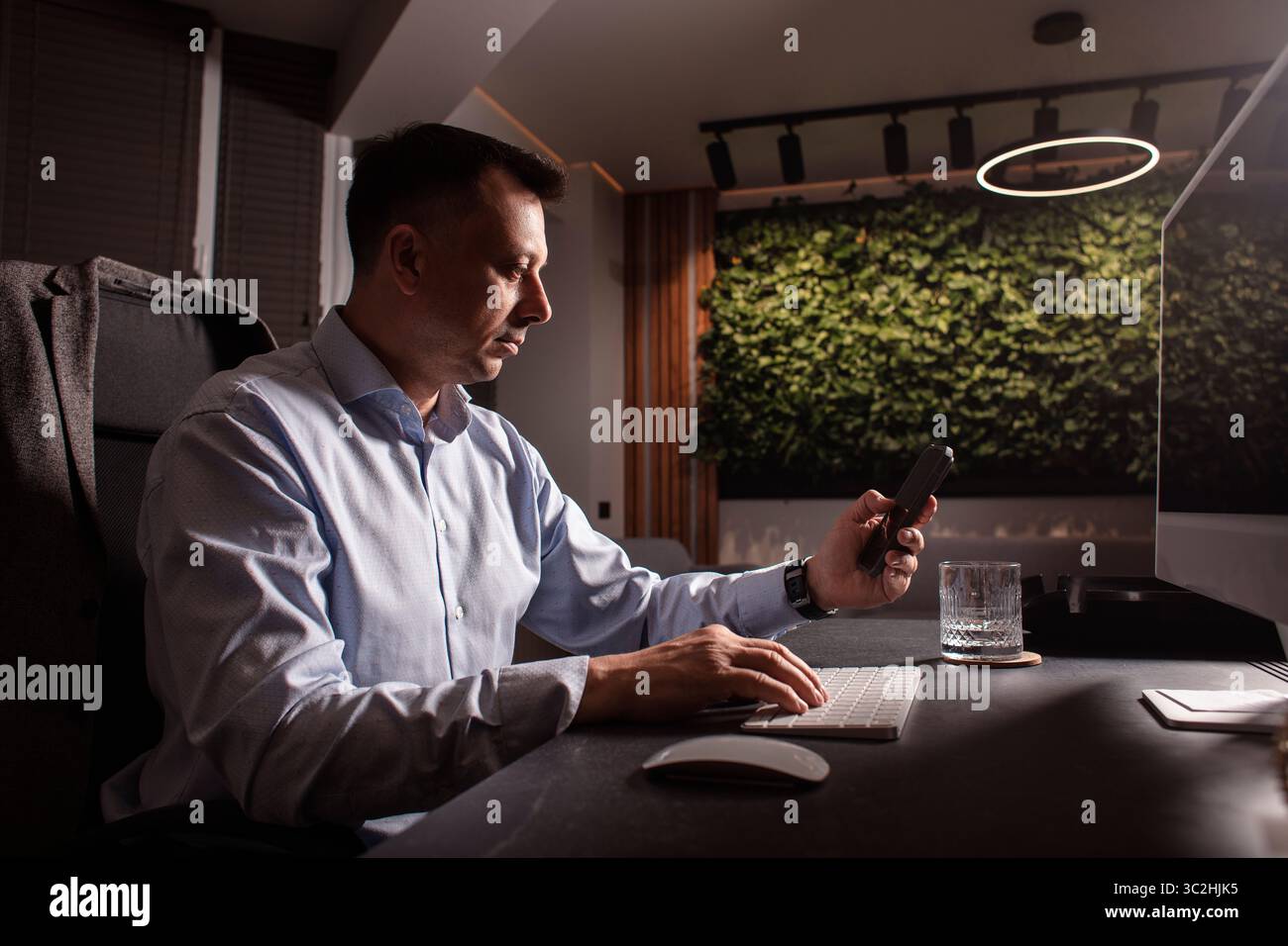 Uomo d'affari con una camicia leggera, tastiera e telefono in un ufficio elegante e buio. Luce da sera, spazio di lavoro elegante, concentrazione sulle attività professionali. Foto Stock