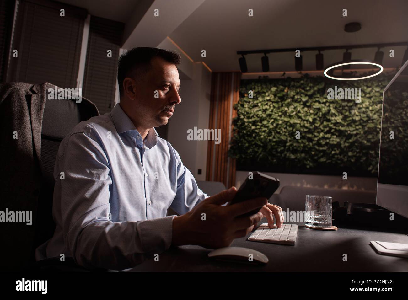 Uomo d'affari con una camicia leggera, tastiera e telefono in un ufficio elegante e buio. Luce da sera, spazio di lavoro elegante, concentrazione sulle attività professionali. Foto Stock