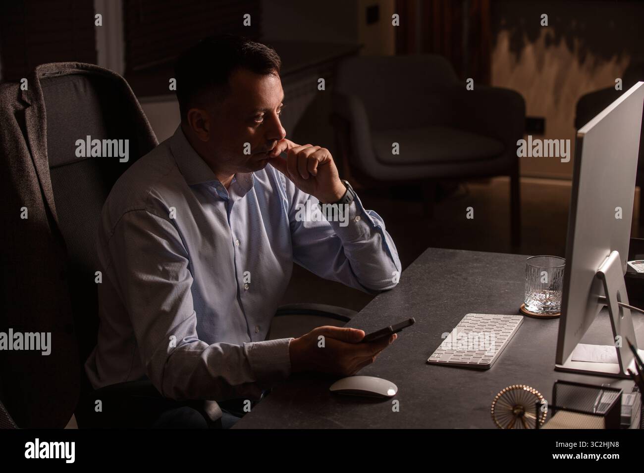 Uomo d'affari con una camicia leggera, tastiera e telefono in un ufficio elegante e buio. Luce da sera, spazio di lavoro elegante, concentrazione sulle attività professionali. Foto Stock
