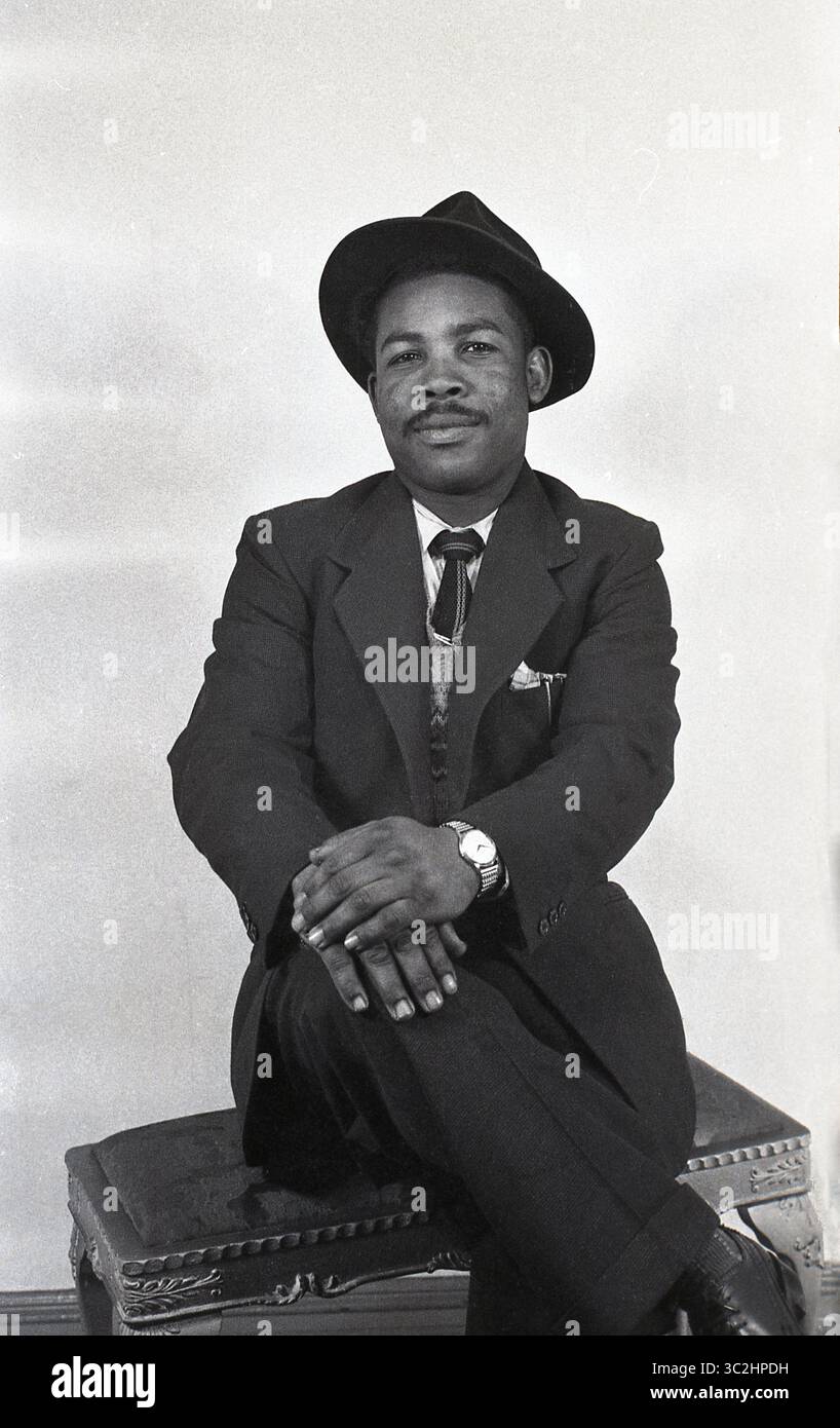 Anni '1950, storico, all'interno di uno studio fotografico... un gentiluomo afro-caraibico vestito con abito e cravatta e cappello seduto su uno sgabello per la sua foto, Inghilterra, Regno Unito. In quest'epoca, avere la tua fotografia scattata in uno studio, in un ambiente formale era popolare, poiché le fotocamere a pellicola da 35 mm e i laboratori fotografici più economici per elaborare la pellicola e fare la stampa non arrivarono fino agli anni '1960 Quindi in quest'epoca, per servizi fotografici di qualità, sei andato da un fotografo professionista in uno studio. Foto Stock