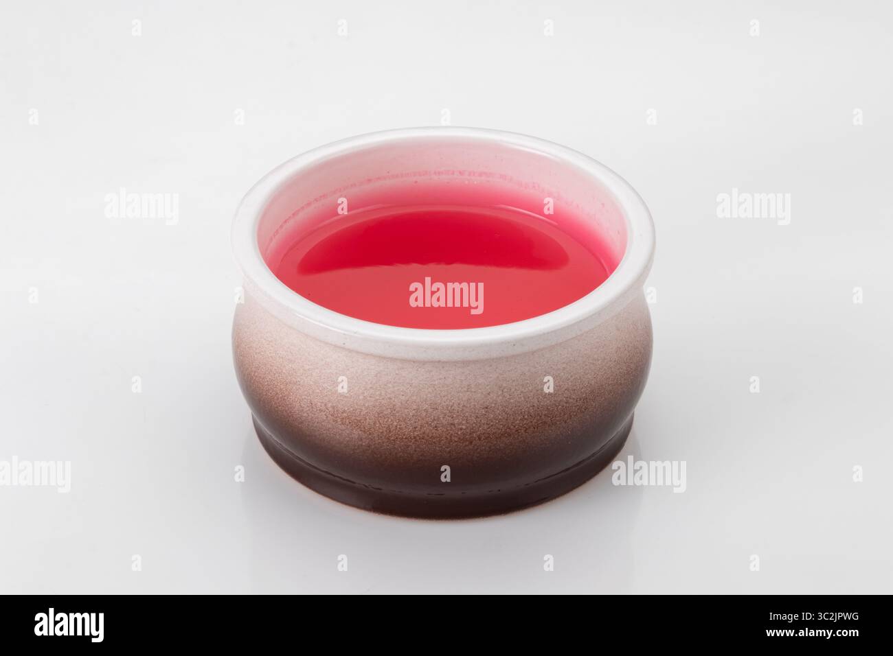 Piccolo recipiente rotondo in ceramica contenente gelatina rossa brillante, isolato su sfondo bianco con riflessi e bokeh Foto Stock