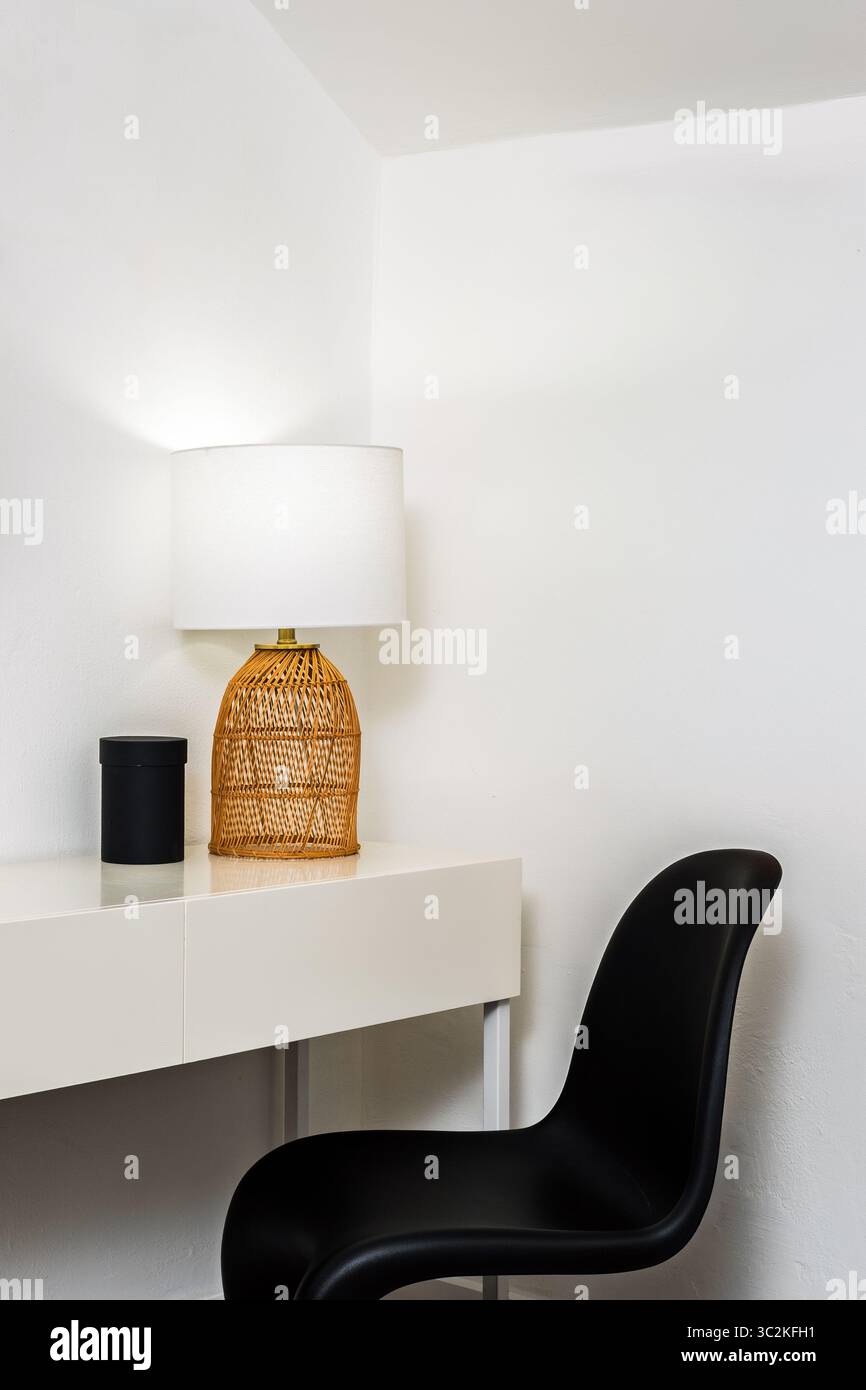 Interni puliti e moderni con una scrivania bianca con una lampada da tavolo in rattan intrecciato e una sedia d'accento nera, adagiata su pareti bianche in uno stile minimalista Foto Stock