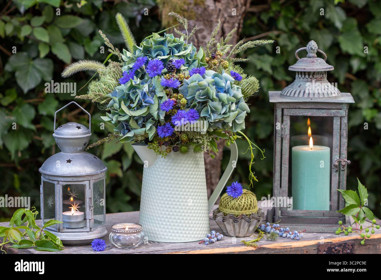 disposizione giardino con bouquet di ortensie blu in fiore, rosa piuma, rami di mora, carote selvatiche e erbe in una caraffa e lanterne vintage Foto Stock