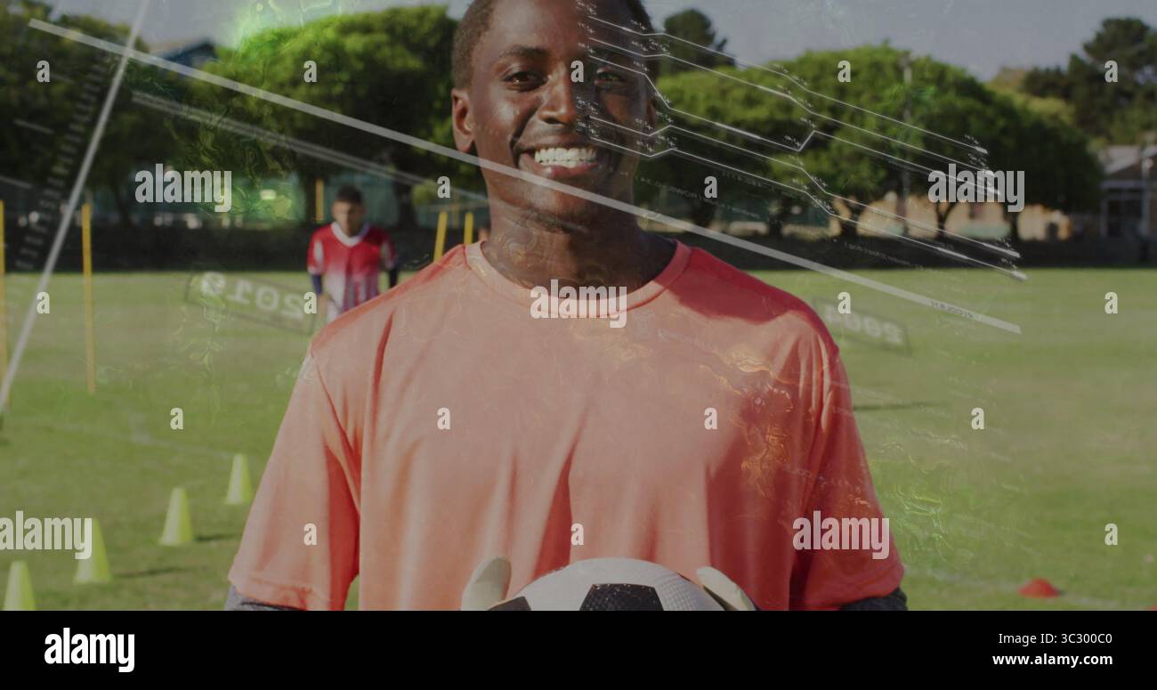 Portiere sorridente che indossa maglia di corallo e guanti che reggono palla da calcio sul campo scolastico, con coni Foto Stock