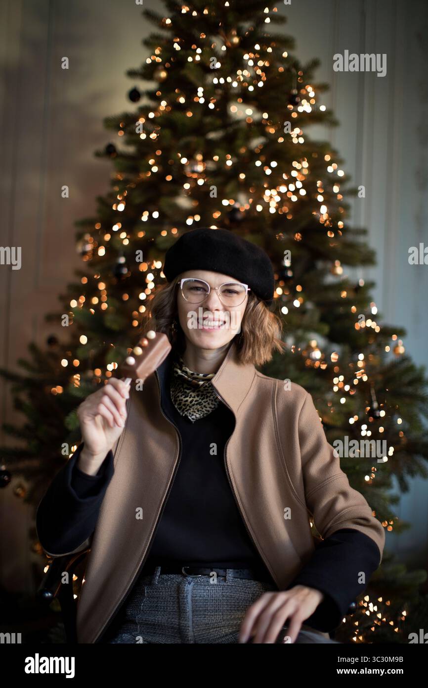 Bella donna in abito invernale seduta davanti all'albero di Natale illuminato, mentre tiene in mano il dolce al cioccolato. Felice donna allegra e alla moda Foto Stock