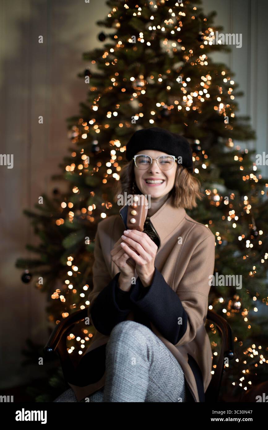 Donna simpatica e allegra in abito invernale seduta davanti all'albero di Natale illuminato, mentre tiene in mano il cioccolato dolce. Donna felice alla moda Foto Stock